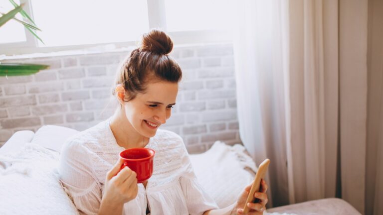 Mulher sorrindo com caneca de café e celular na cama pela manhã